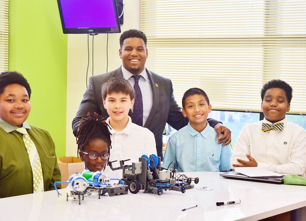 A UVA undergraduate student poses with five school children at an after-school robotics class, with some of their work in the foreground.