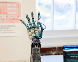 A robotic hand sits on a table in front of a poster and computer screen.