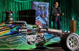 Behl speaks next to a screen showing Cavalier Autonomous Racing celebrating its recent win. An Indy Autonomous Challenge car sits in the foreground.