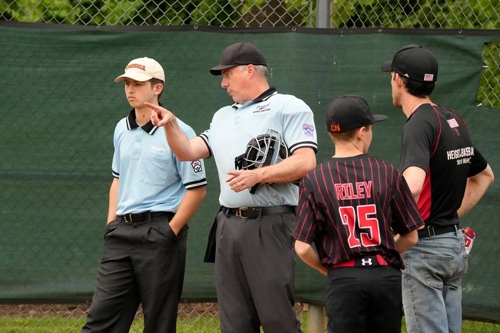 A youth league baseball umpire explains to another umpire while a player and another adult look on.