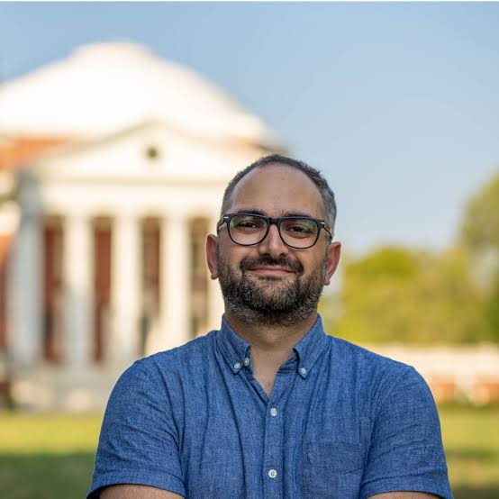 Arsalan Heydarian stands outdoors wearing glasses and a short-sleeved blue button-up shirt, with arms crossed. A columned building is softly blurred in the background, surrounded by green lawn and trees under a clear sky.
