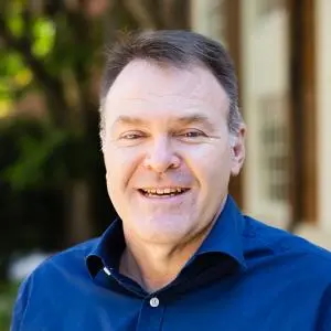 Brian Smith in a professional headshot, wearing a navy blue collared shirt and facing the camera outdoors, with a softly blurred building and greenery in the background.