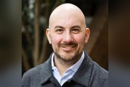 Daniel Otero-Leon head-and-shoulders portrait outdoors, wearing a light blue collared shirt under a dark gray jacket. Daniel Otero-Leon faces the camera against a softly blurred background of trees and warm-toned foliage.