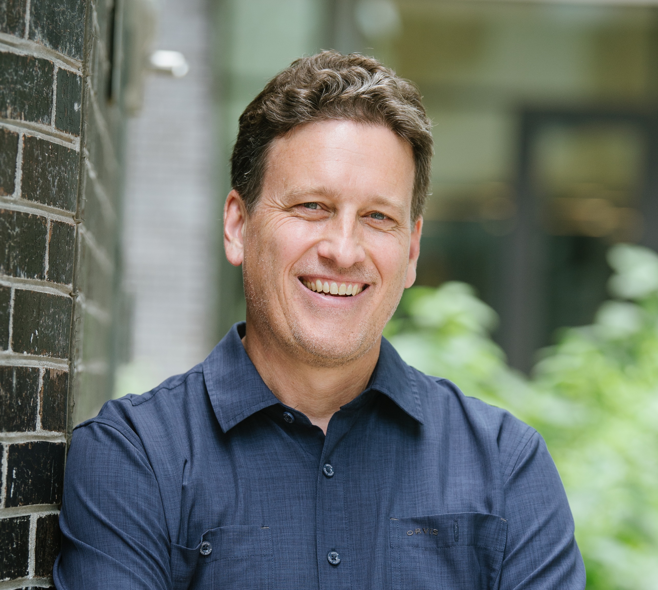 David Luebke, Vice President at NVIDIA Research, stands beside a brick wall wearing a blue button-down shirt. The professional headshot is taken indoors with a softly blurred background of windows and greenery.
