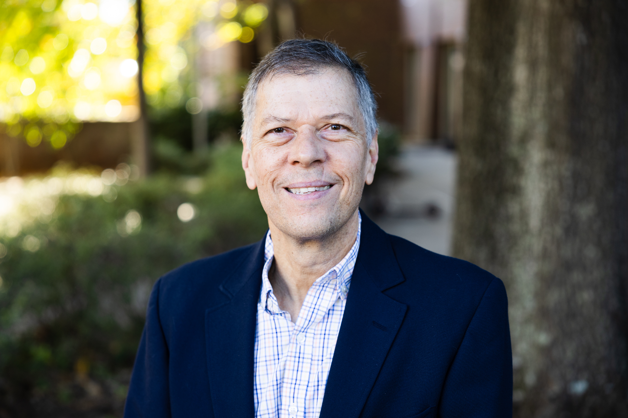 Eric Loth poses outdoors in a head-and-shoulders portrait, wearing a navy blazer over a light checkered button-down shirt. Trees, greenery, and building columns appear softly blurred in the background.