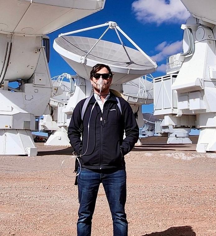 Michael standing outside by satellite dishes wearing oxygen mask and hat