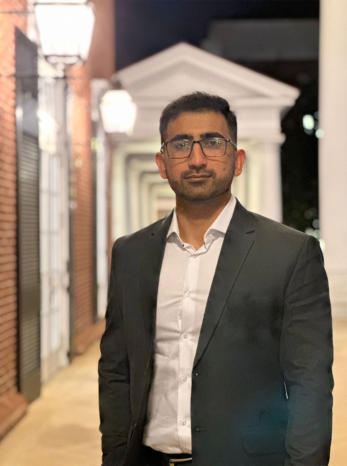A man wearing glasses and a suit stands in an outdoor corridor with brick columns and white archways at night.