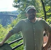 man wearing tan shirt and glasses standing outside with view of beautiful bridge