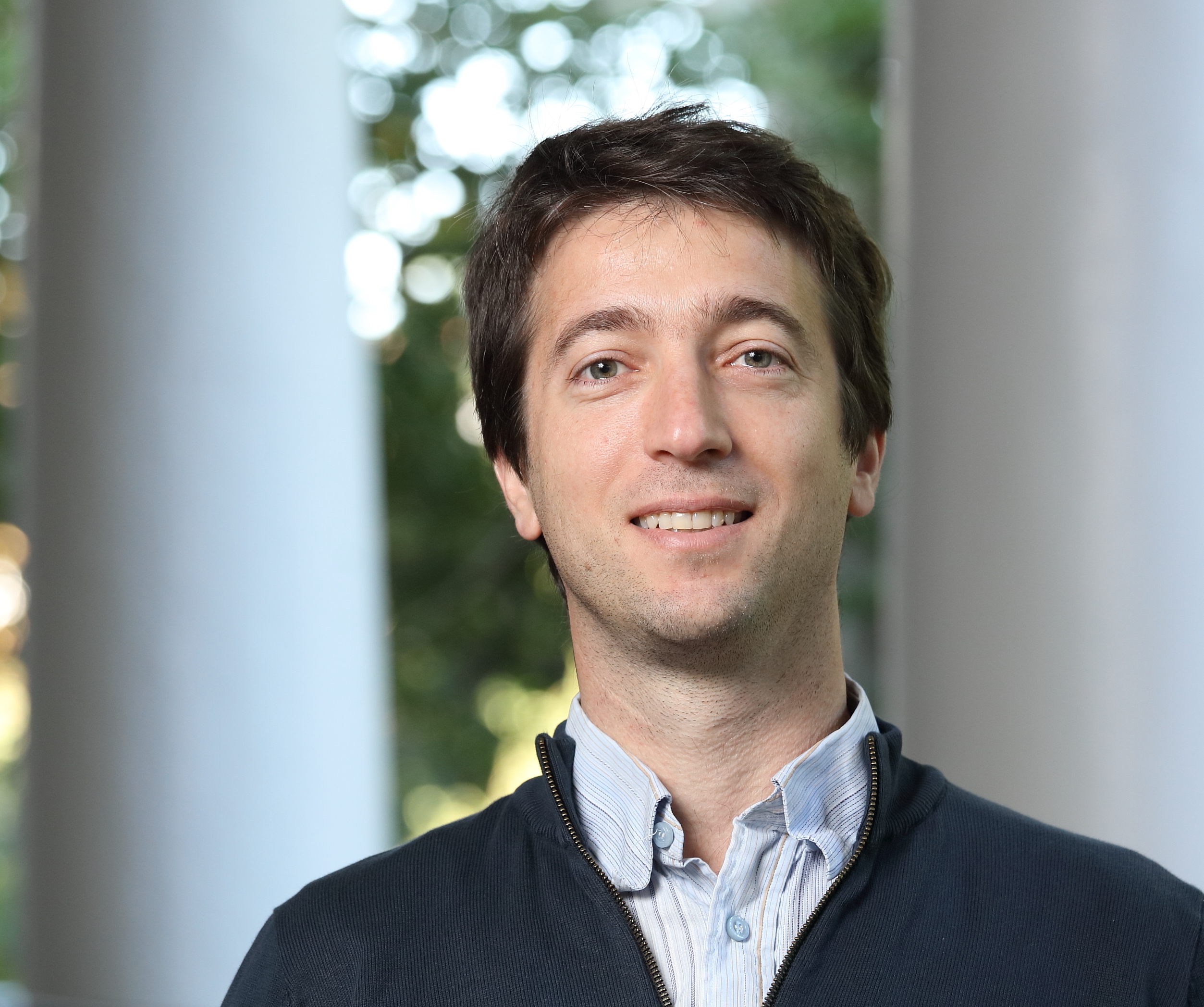 Sebastian Elbaum poses for a head-and-shoulders portrait indoors, wearing a light striped button-down shirt under a dark quarter-zip sweater. Blurred windows and greenery in the background create a softly lit setting.