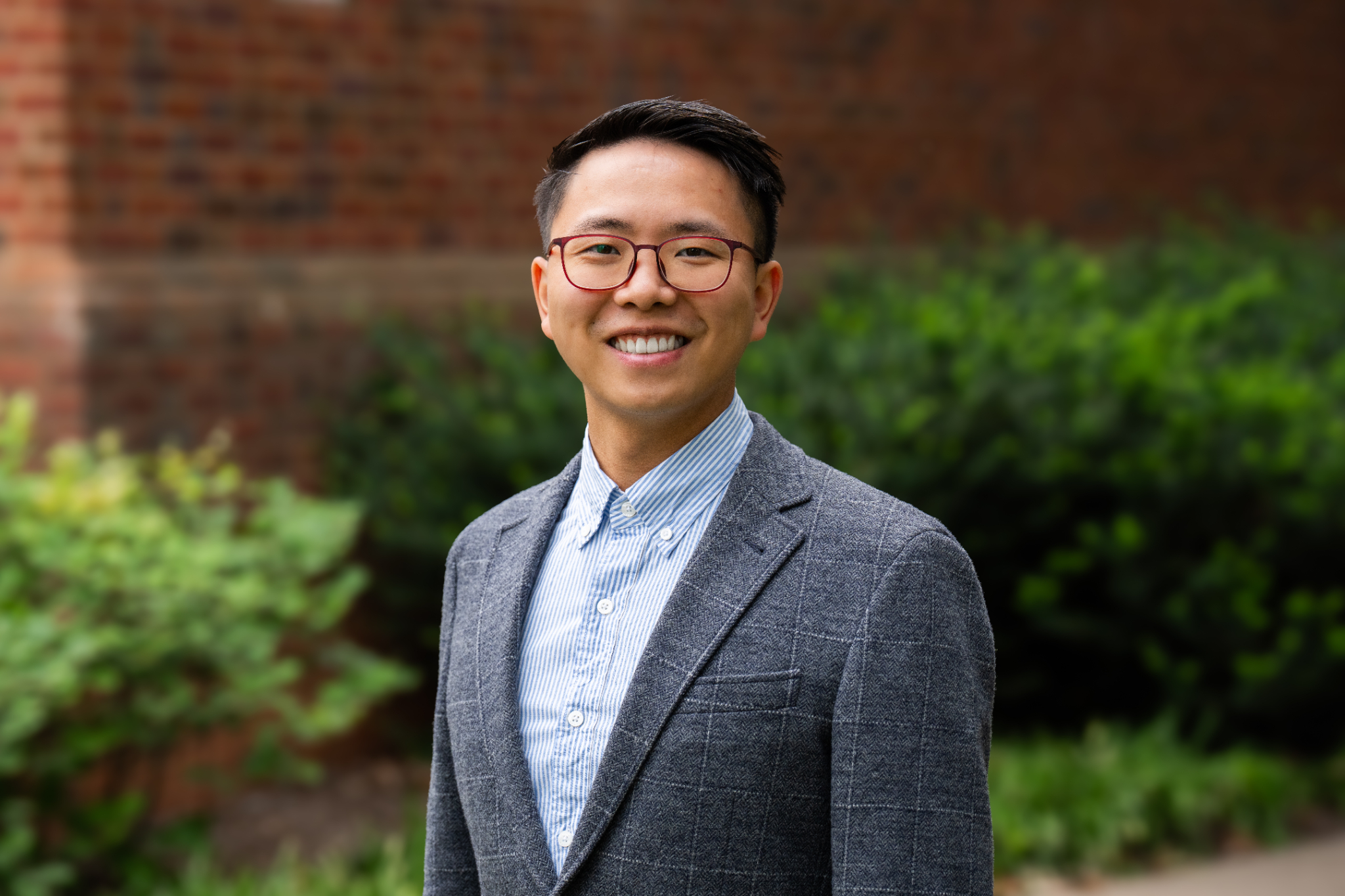 Xun Zhao stands outdoors in front of a softly blurred brick building and greenery, wearing red-framed glasses, a light blue striped button-down shirt, and a gray checkered blazer while facing the camera.