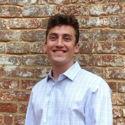Head-and-shoulders portrait of a person standing in front of a textured red brick wall. The person is facing the camera and wearing a light-colored, checkered button-down shirt. The image is evenly lit and framed from the chest up, with the brick wall filling the background.