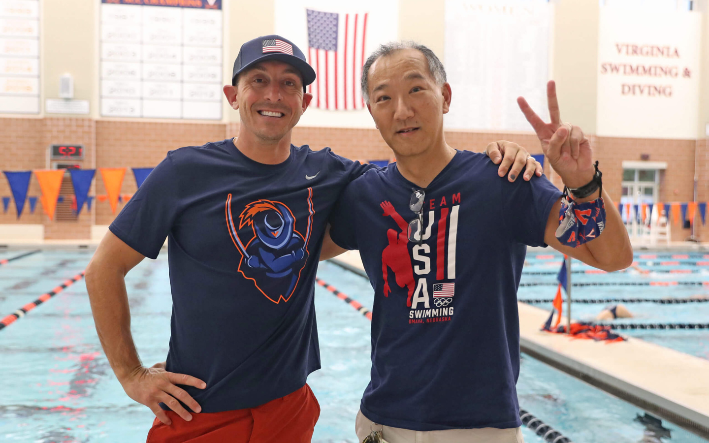 Two men wearing T-shirts posing in front of a competitive swimming pool, one giving the victory sign.