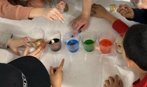 Children gather around cups of liquid in different colors.