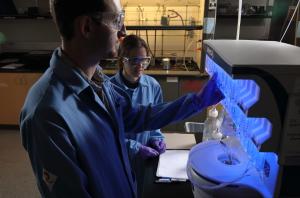 A chemical engineering professor and her student work at a peptide synthesizing machine that glows blue in a darkened lab.