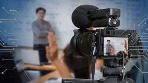 A video camera viewfinder in the foreground captures a teacher at the front of a classroom where several students have their hands raised.