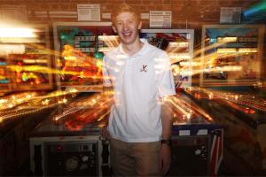 A smiling young man standing in front of a bank of brightly lit, blurred pinball machines.