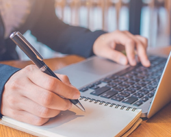 student typing on laptop and taking notes with pen and pad