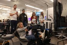 Woman sits in a racecar seat for a full size video gam rig in the lab surrounded by 4 others smiling and watching her