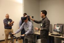 Man adjusts VR headset on another man sitting on a stationary bike with other look on