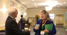 A man stands in a hallway speaking to a woman as they hold cups of coffee