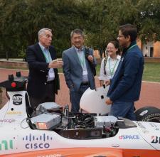 4 people stand around autonomous racecar with the hood removed to reveal the inner parts