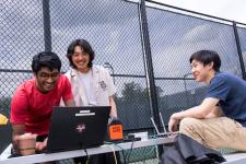 A group of people looking at a computer on a table set up outside.