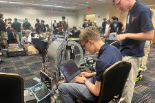 Two students working on a computer with a group of people in the background.