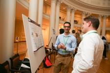 Two attendees discuss a research poster during UVA Link Lab Research Day, holding refreshments in a networking area with columns in the background.