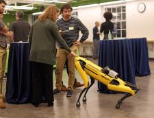 Woman pets Robot Dog "Spot" who is in a kneeling pose next to a man standing looking on at reception