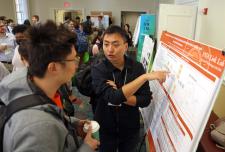 Man stands pointing to a poster in a reception in front of another man speaking surrounded in a room with other posters and people talking