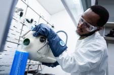 A man wearing a white lab coat, safety glasses and blue gloves pours liquid nitrogen into a flask. 