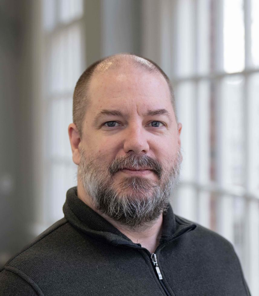 White man with buzzed hair and a salt-and-pepper beard wearing a dark grey half-zip sweatshirt stands in front of a bank of windows.