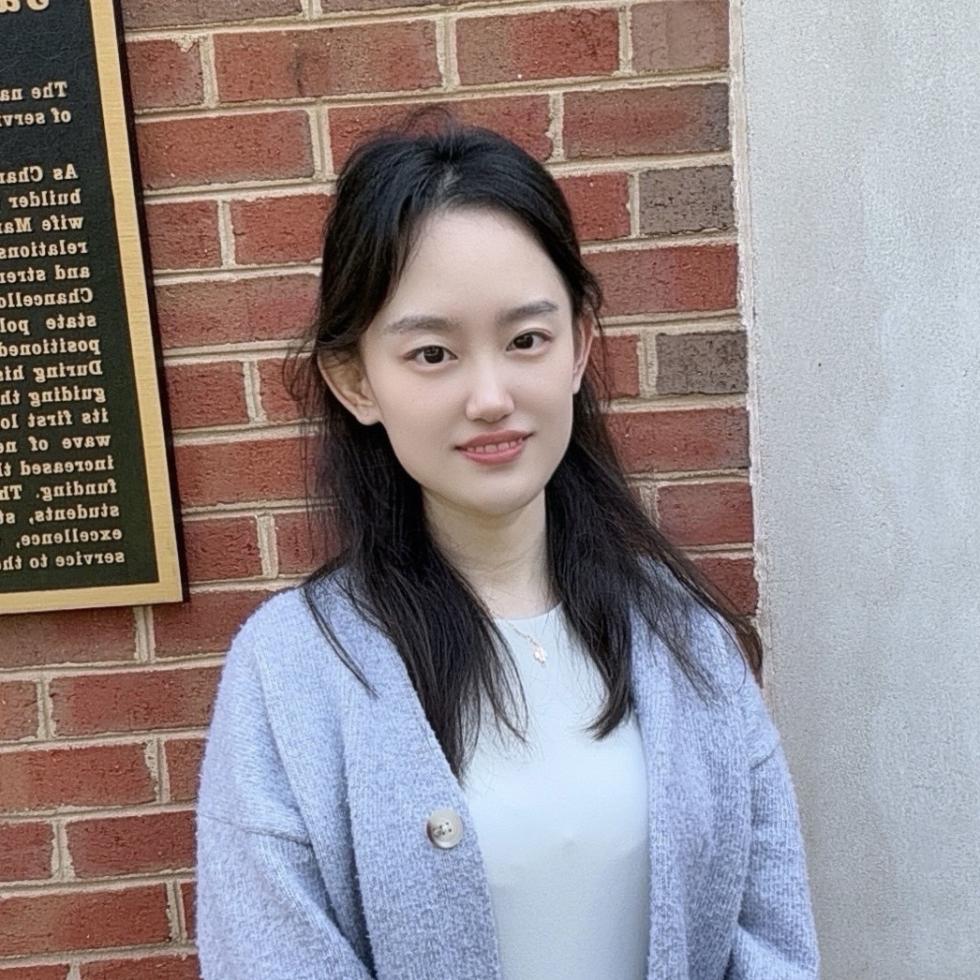 Image of woman in grey cardigan sweater standing in front of a brick wall smiling
