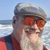 man with beard wearing red shirt, sun glasses, and beret smiling in front of an ocean