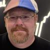 man wearing black shirt, glasses, and mutli-colored cap smiling