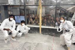 A group of people wearing white protective gear in the foreground with a bunch of smiling students behind a transparent barrier behind them.