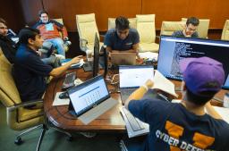 UVA students seated around a conference room table covered with computers, computer monitors, and paper and pencils, work intently during the qualifying round  of the Mid-Atlantic Collegiate Competition.